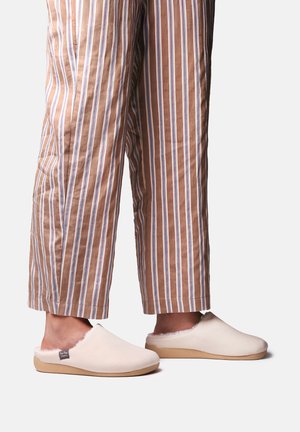 Person iført beige og hvide stribede bukser og cremefarvede slip-on tøfler med lysebrune såler står på en ensfarvet baggrund.