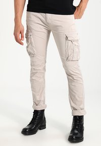 Beige cargobroek met meerdere zijzakken, opgerolde broekspijpen en een gladde textuur, gecombineerd met zwarte veterschoenen.