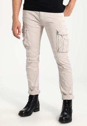 Pantalons cargo beiges avec plusieurs poches latérales, ourlets retroussés et texture lisse, associés à des bottes noires à lacets.