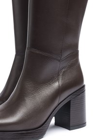Botas de tobillo de cuero marrón con un acabado brillante, punta cuadrada y tacón grueso y texturizado. Presenta sutiles acentos de costura.