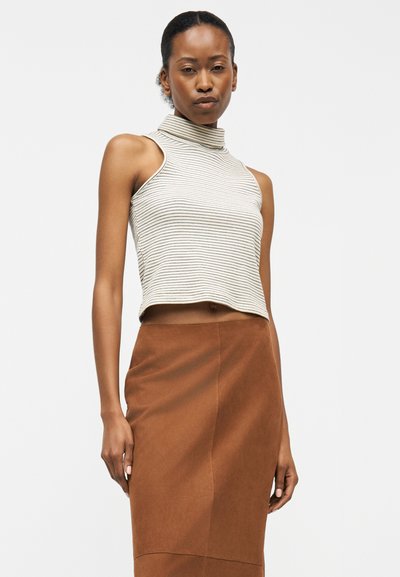 Γυναίκα που φοράει αμάνικη ριγέ τοπ με ψηλό λαιμό και φούστα καφέ, ψηλόμεση, στέκεται μπροστά σε απλό φόντο.