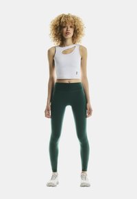 Mujer con cabello rizado está de pie de frente, vistiendo una camiseta sin mangas blanca recortada con cortes y unas mallas de talle alto verde oscuro, además de zapatillas blancas.