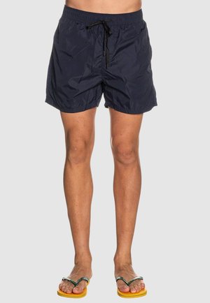 Uomo che indossa pantaloncini da bagno blu navy e infradito gialli con cinturini verdi, in piedi contro uno sfondo grigio chiaro uniforme.