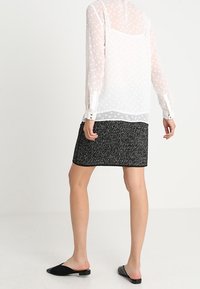 Průhledná bílá blůza s dlouhými rukávy a puntíky, doplněná černou texturovanou mini sukní. Černé slip-on boty dokončují celý outfit.