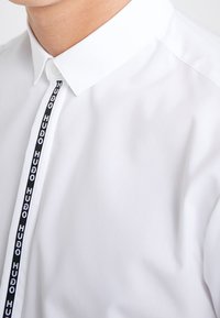 Valkoinen paita, jossa on perinteinen kaulus ja pystysuora musta nauha, jossa on toistuvasti sana "HUGO". Tasainen kangas, jossa on vähäinen rakenne.