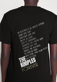 Camiseta negra con texto en blanco y dorado metálico en la parte trasera, mostrando el nombre de la marca y el eslogan en diferentes fuentes y orientaciones.