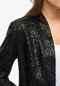 Zwart jasje met pailletten en een gestructureerd oppervlak, open model zonder sluiting. Heeft een losse pasvorm en lange, licht uitlopende mouwen.