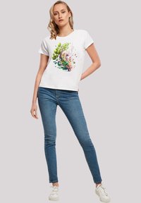 White t-shirt featuring a colorful floral design with green leaves and pink blossoms, paired with blue jeans and white sneakers.