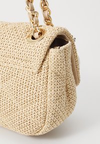 Beige vevd toteveske med en teksturert overflate, med en gullfarget kjedestropp og en subtil åpning på toppen.