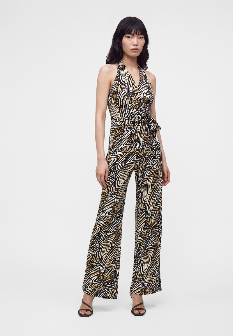 Femme portant une combinaison sans manches à col halter avec un motif tourbillon abstrait noir, blanc et marron et des talons ouverts noirs.