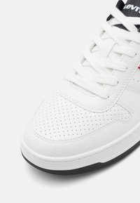 Vita lädersneaker med perforerat tåkåpa, platta vita snören och svart sula. Har en röd och svart logotyp på tungan.