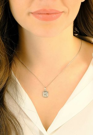 Collana d'argento con un ciondolo rettangolare che presenta un'incisione dettagliata, indossata su una catena sottile sopra una camicetta bianca.