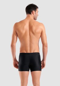 Schwarze Badeshorts mit niedriger Taille, aus glattem Stoff gefertigt, mit einfachem Design und engem Sitz, von hinten betrachtet.
