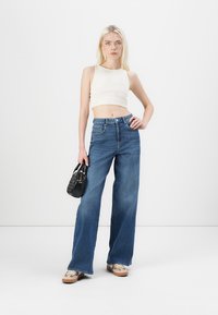 Jeans din denim albastru, cu pantaloni largi și talie înaltă, asortați cu un top alb ajustat. Accesorii includ o geantă de umăr neagră și pantofi cu un model.