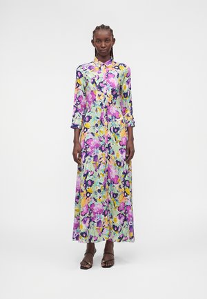 Γυναίκα όρθια με φόρεμα μακρυμάνικο, μακρύ που κουμπώνει μπροστά με πολύχρωμο λουλουδάτο μοτίβο και μαύρες ψηλοτάκουνες πέδιλα με λουριά.