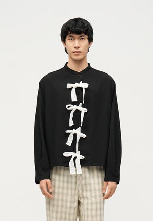 Νεαρός άνδρας που φορεί μαύρο πουκάμισο με μακριά μανίκια, στο μπροστινό μέρος του οποίου υπάρχουν τέσσερα λευκά δαντελωτά φιογκάκια, και ανοιχτόχρωμο καρό παντελόνι, στέκεται μπροστά σε απλό φόντο.