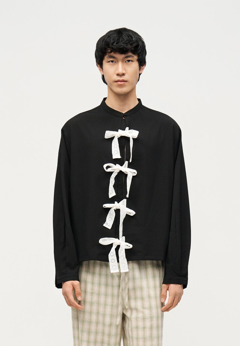 Νεαρός άνδρας που φορεί μαύρο πουκάμισο με μακριά μανίκια, στο μπροστινό μέρος του οποίου υπάρχουν τέσσερα λευκά δαντελωτά φιογκάκια, και ανοιχτόχρωμο καρό παντελόνι, στέκεται μπροστά σε απλό φόντο.