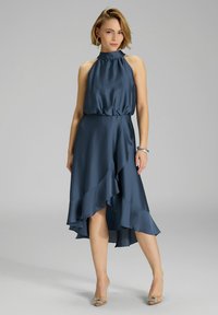 Robe sans manches à col halter en tissu bleu foncé lisse, avec un ourlet à volants asymétrique. Associée à des talons beige embellis.