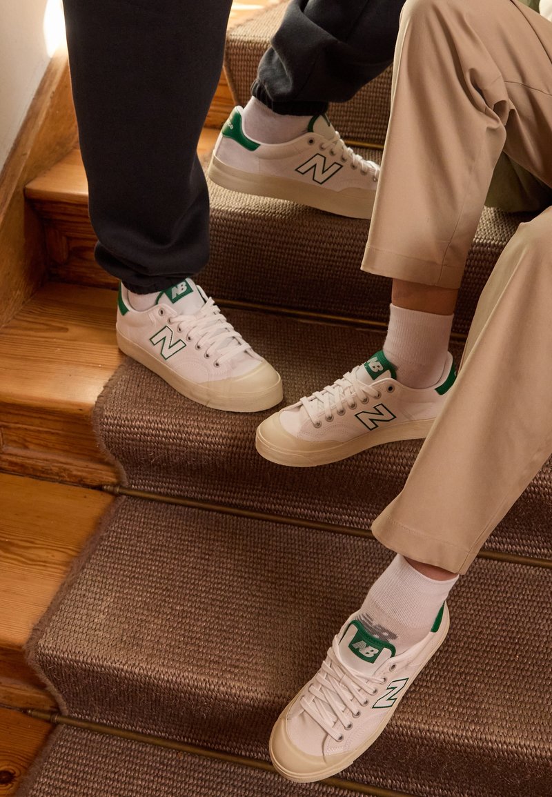 Deux personnes portant des baskets New Balance blanches et vertes, assises et debout sur des escaliers en bois recouverts de moquette.