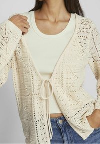 Cárdigan de ganchillo crema con abertura frontal, patrones geométricos intrincados y cierre de lazo. Llevado sobre una camiseta sin mangas de color claro.