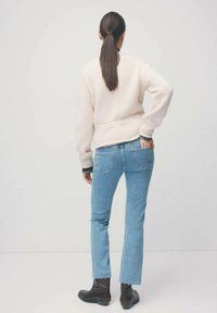 Kobieta z długimi ciemnymi włosami związanymi w kucyk, ubrana w kremowy sweter, jasnoniebieskie dżinsy i czarne botki, stojąca z jedną ręką w kieszeni.