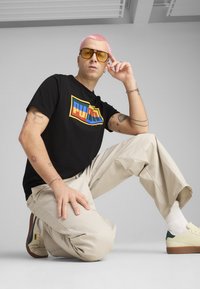 Melna t-krekla ar daudzkrāsainu "PUMA" logotipu, bēšas krāsas bikses un krēmkrāsas sporta apavus ar zaļiem akcentiem. Siluetam ir rozā mati un dzelteni saulesbrilles.