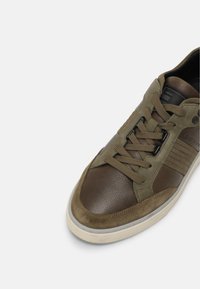 Brown and olive green sneaker with leather and suede materials, featuring textured panels, six eyelets, and a rubber sole.