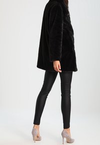 Svart faux fur-kappa med en avslappnad passform, breda ärmar och en mjuk textur, kombinerad med svarta stretchbyxor och ljusgrå klackar.