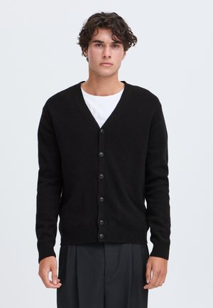 Jeune homme aux cheveux bouclés portant un cardigan noir boutonné sur une chemise blanche et un pantalon sombre, debout devant un fond clair uni.