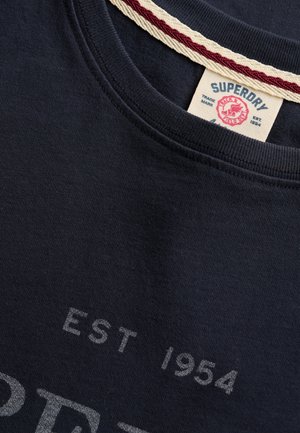 Nærbillede af mørk navy Superdry-skjorte med hvid og rød nakkebånd og delvist "EST 1954" tekst på stoffet.
