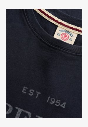 Close-up van de donkerblauwe Superdry-overhemdkraag met witte en rode nektape en gedeeltelijke tekst "EST 1954" op de stof.
