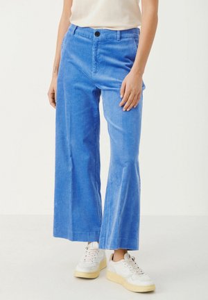 Persoon draagt blauwe corduroy broek met wijde pijpen, gecombineerd met witte sneakers en een wit mouwloos topje, staand tegen een effen achtergrond.