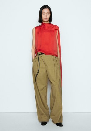 Personne portant un haut rouge asymétrique sans manches et un pantalon ample taille haute kaki avec un détail de lien noir, debout devant un fond clair uni.