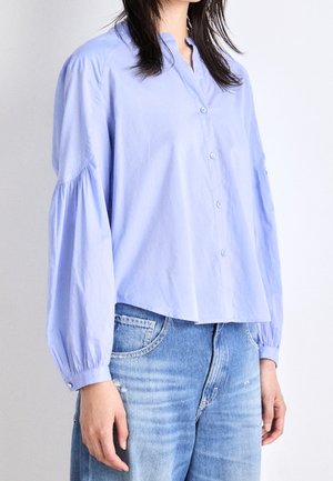 Femme portant un chemisier bleu clair boutonné avec des manches longues bouffantes et un jean bleu clair sur un fond uni.