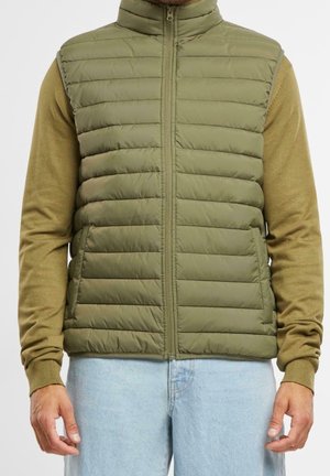 Mann mit olivgrüner gesteppter Weste über einem hellbraunen Langarmhemd und hellblauen Jeans, steht mit entspannten Händen seitlich am Körper.