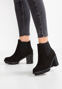 Beine in schwarzen Wildleder-Ankle Boots mit Blockabsätzen und Profilsohlen, kombiniert mit dunkelgrauen Skinny Jeans mit fransigem Saum.