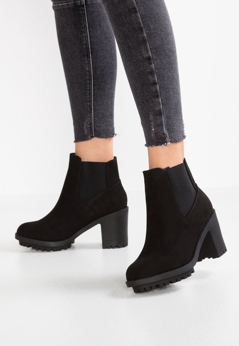 Beine in schwarzen Wildleder-Ankle Boots mit Blockabsätzen und Profilsohlen, kombiniert mit dunkelgrauen Skinny Jeans mit fransigem Saum.