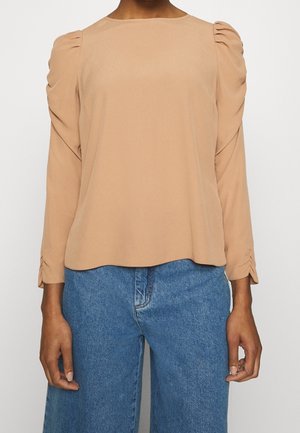 Personne portant un chemisier beige à manches longues avec des épaules froncées et un jean bleu taille haute sur un fond uni.