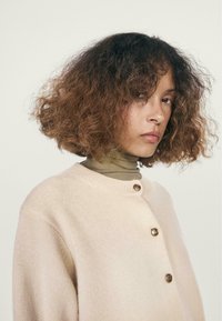 Cardigan a maglia in un morbido beige con scollo rotondo, caratterizzato da tre bottoni color oro e una finitura testurizzata. Abbinato a un dolcevita verde.