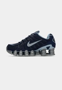 Tumši zili un sudraba sporta apavi ar segmentētu mīkstinātu zoli un sudraba Nike swoosh sānos, attēloti labajā profilā uz baltas fona.