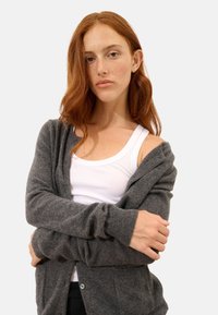 Szary kardigan o miękkiej teksturze, z długimi rękawami i zapięciem na guziki. Noszony na białym topie na ramiączkach, podkreślając stylizację warstwową.