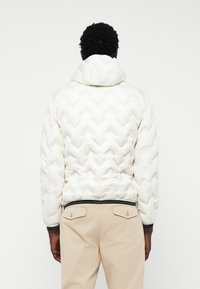 Witte pufferjack met een gestructureerd, chevronpatroon en een capuchon. Voorzien van een zwarte elastische tailleband en zwarte accenten bij de manchetten.