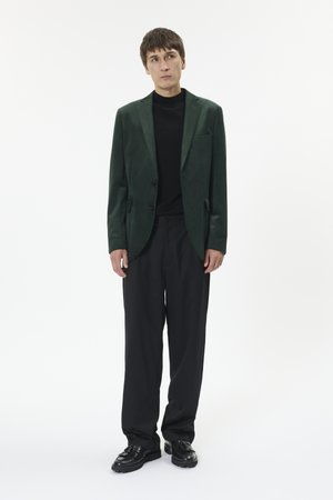 Młody mężczyzna w ciemnozielonym blezerze, czarnym golfie, luźnych czarnych spodniach i czarnych mokasynach z chwostami, stojący na białym tle.