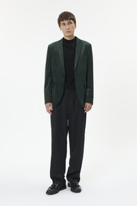 Jaunas vyras, dėvintis tamsiai žalią švarką, juodą golfą, laisvas juodas kelnes ir juodus batelius su kutais, stovintis priešais baltą foną.