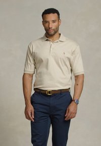 Polo bej cu un decolteu cu trei butoane și logo brodat pe piept, asociat cu pantaloni negri închis. Textură fină din amestec de bumbac.