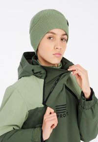 Beanie verde a maglia con texture a coste, abbinato a un giubbotto verde con colletto alto, dotato di chiusura a zip e logo nero.