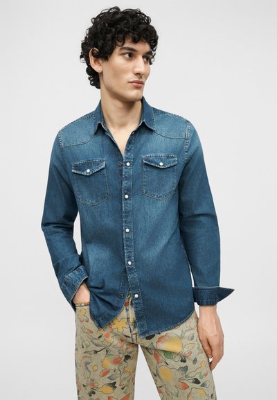 Jeune homme aux cheveux bouclés portant une chemise en jean boutonnée et un pantalon à motif floral, debout avec une main dans la poche, regardant sur le côté.