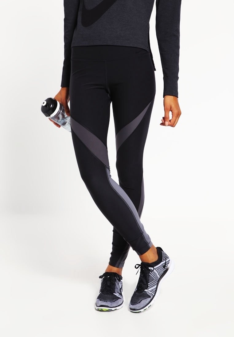 Personne portant des leggings de sport noirs et gris, des chaussures de course noires à motifs, tenant une bouteille d'eau transparente sur un fond blanc.
