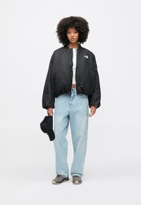 Czarna kurtka bomberka z zamkiem z przodu, o luźnym kroju, z logo na piersi; jasnoniebieskie szerokie dżinsy; szare buty sportowe; czarna czapka trzymana w ręce.