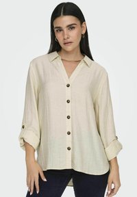 Beige overhemd met knopen en een gestructureerde afwerking, voorzien van een kraag, opgerolde mouwen en donkere knoopaccenten.